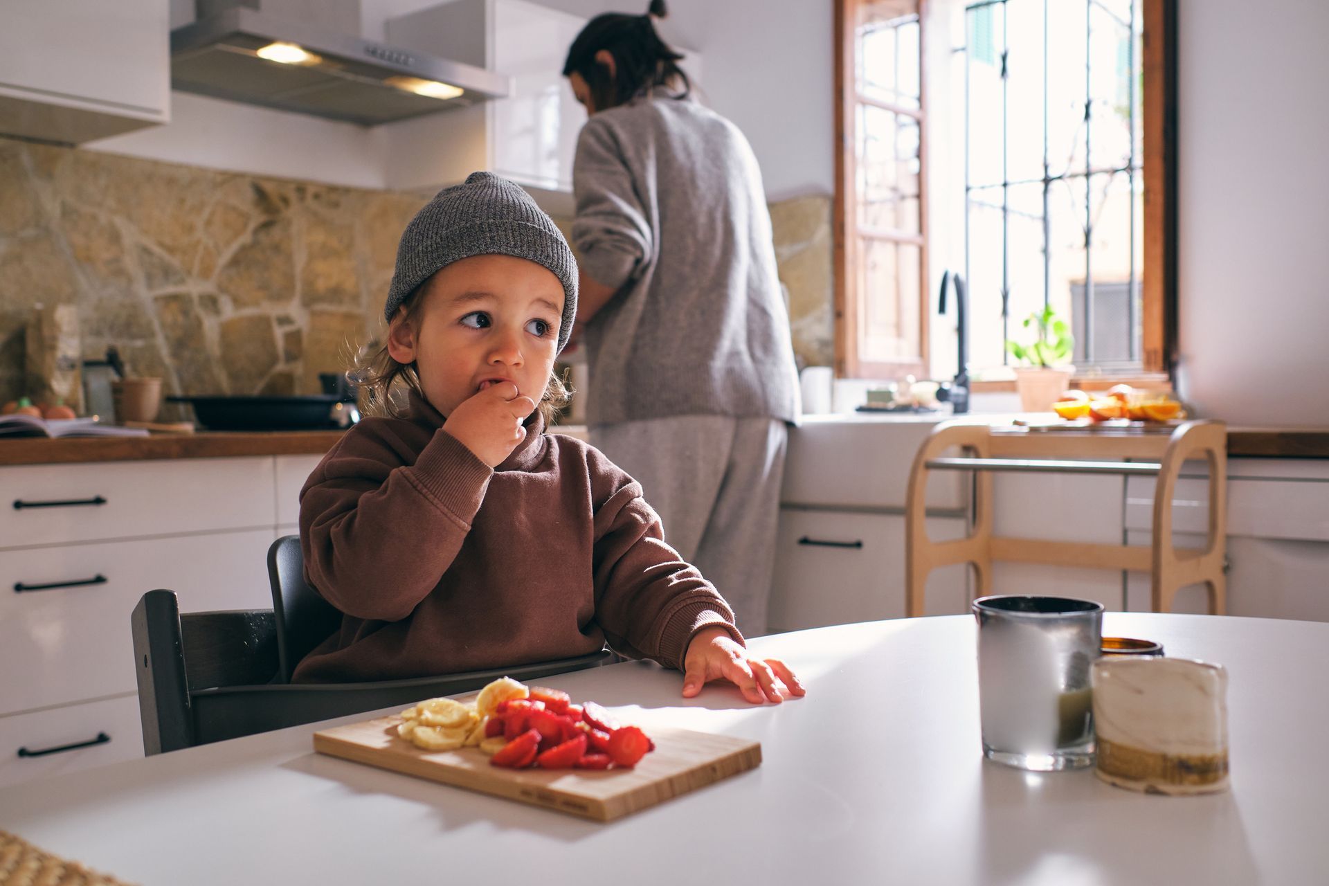 Child in a highchair eating fruit at a kitchen table; a person stands at a sink in the background.