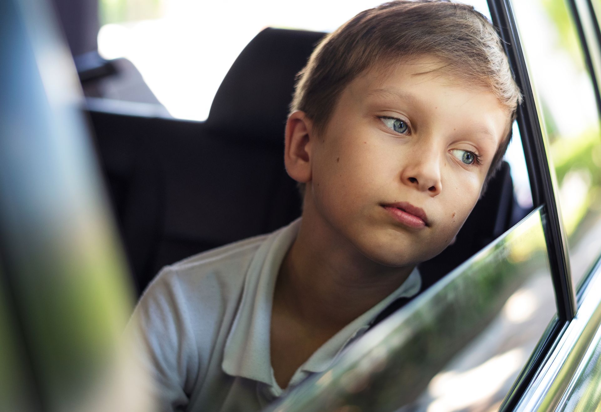 Boy looks out car window, appearing thoughtful, light blue eyes, white shirt, indoor setting.