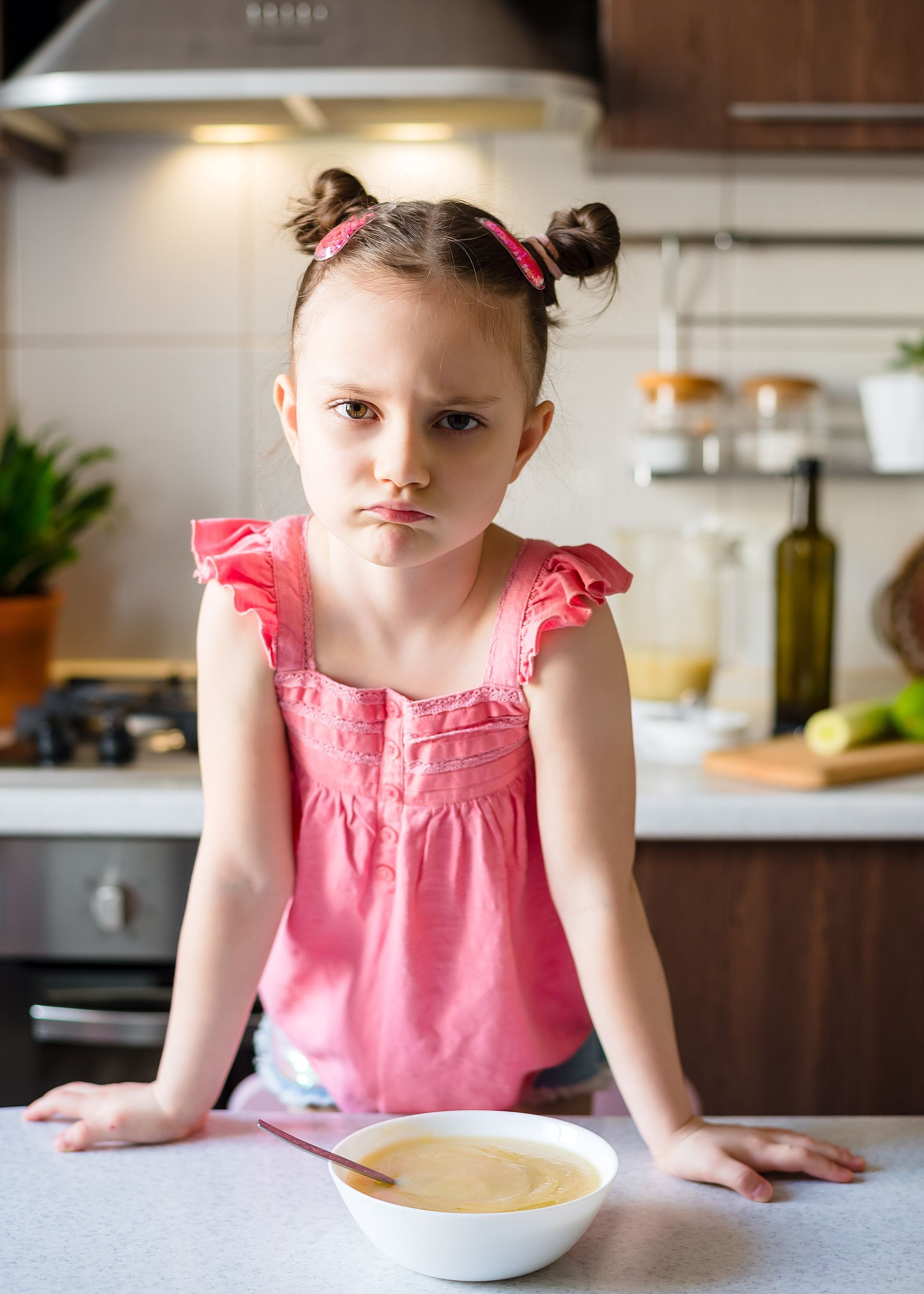 Girl with pigtails scowls at a bowl of food, leaning on a counter in a kitchen.