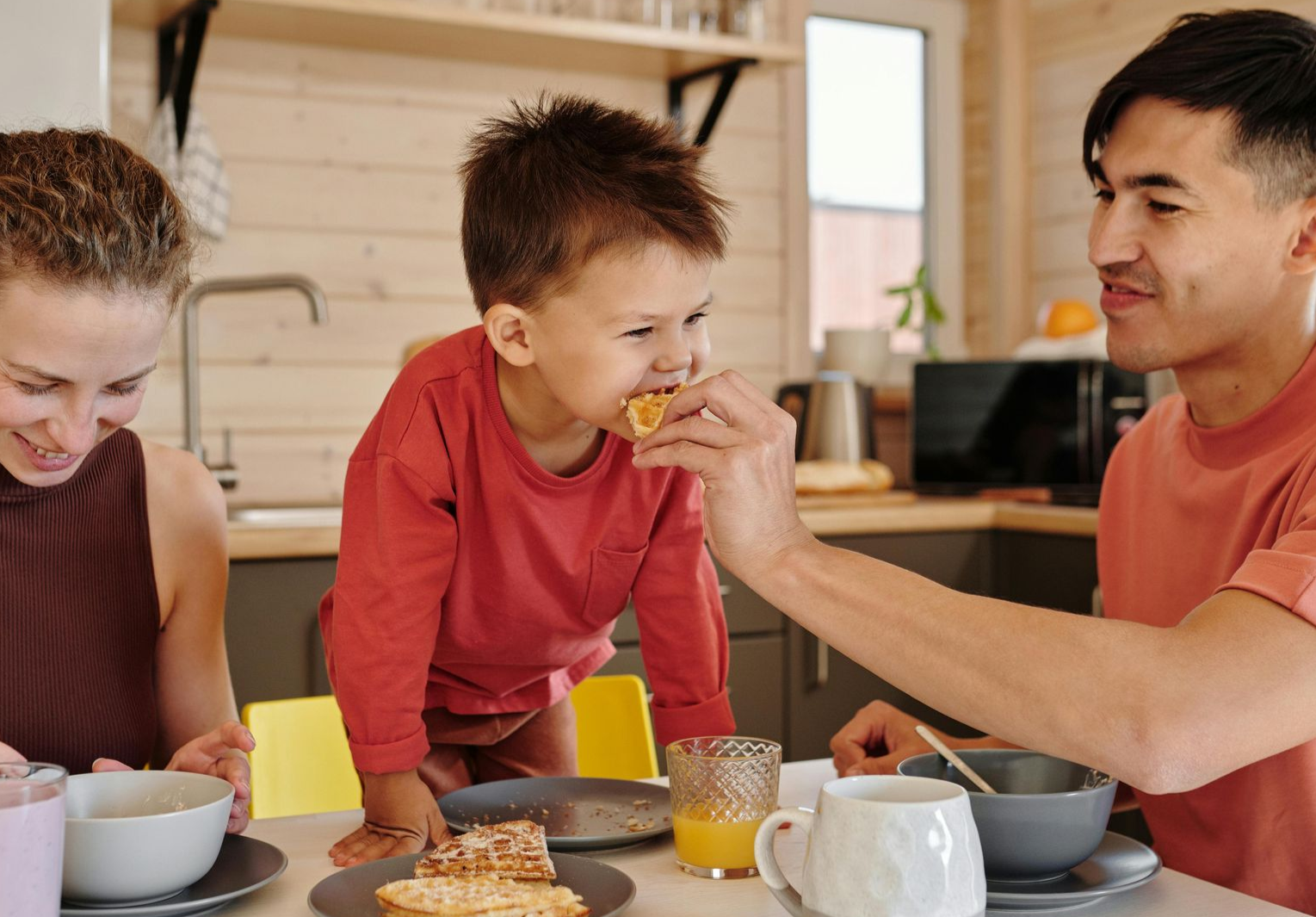 Family in kitchen, child eating food from father, mother at table.