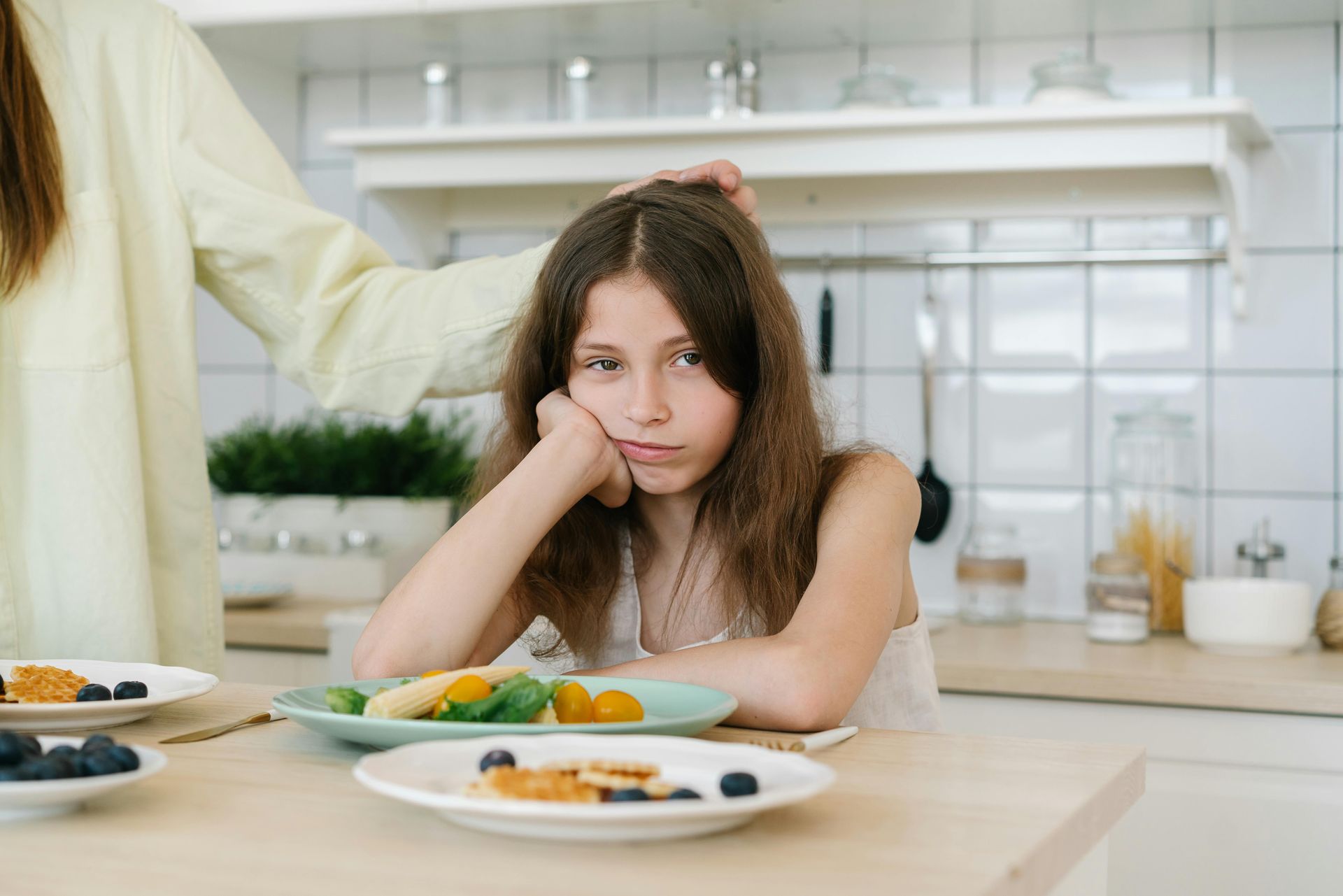 Girl looking sad at breakfast table, hand on cheek, parent's hand on her head.