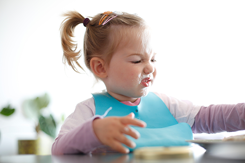 Toddler with a disgusted expression, wearing a bib, sits at a table with food on face.