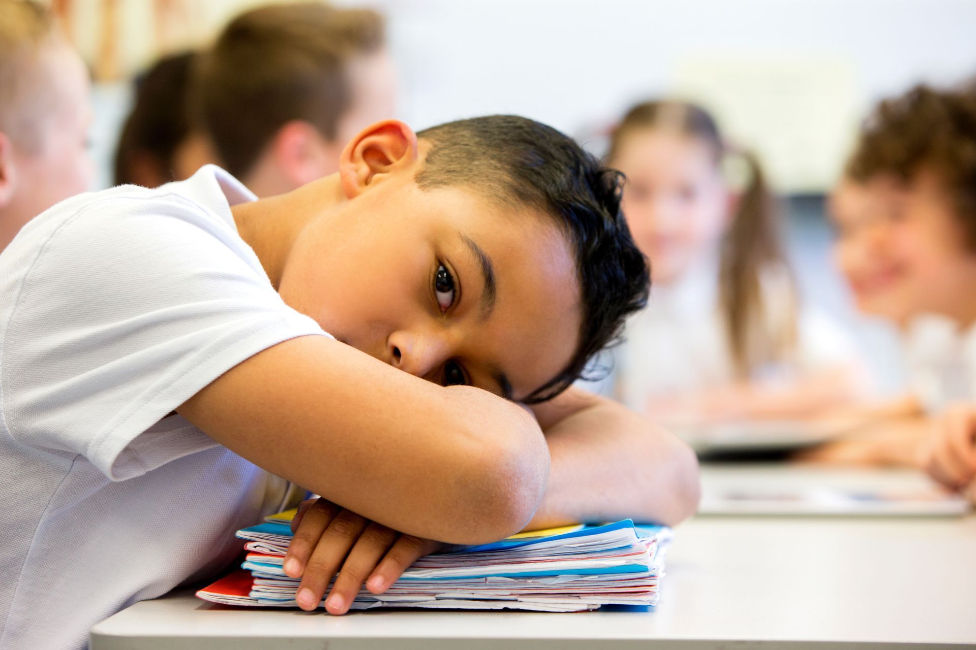 Boy resting head on a stack of papers at a desk in a classroom, looking tired.