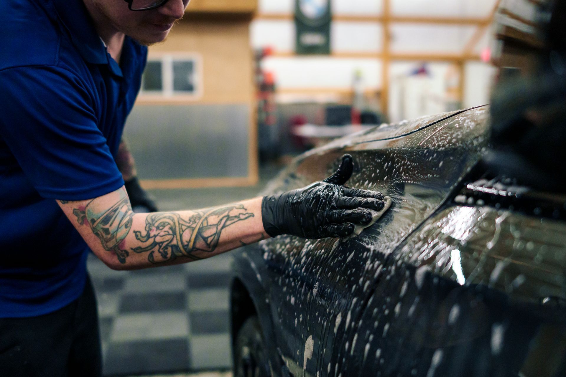 Person washing a black car with soapy water, wearing gloves and a blue shirt.