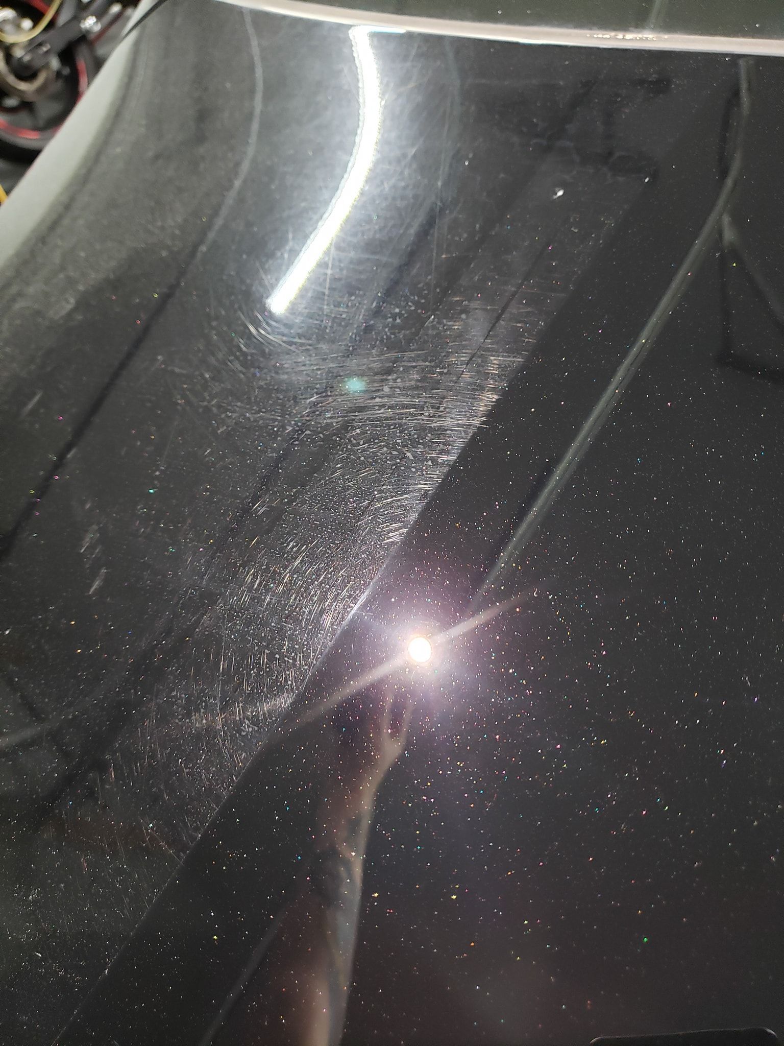 Black car hood with swirl marks and speckles, reflecting a bright light.