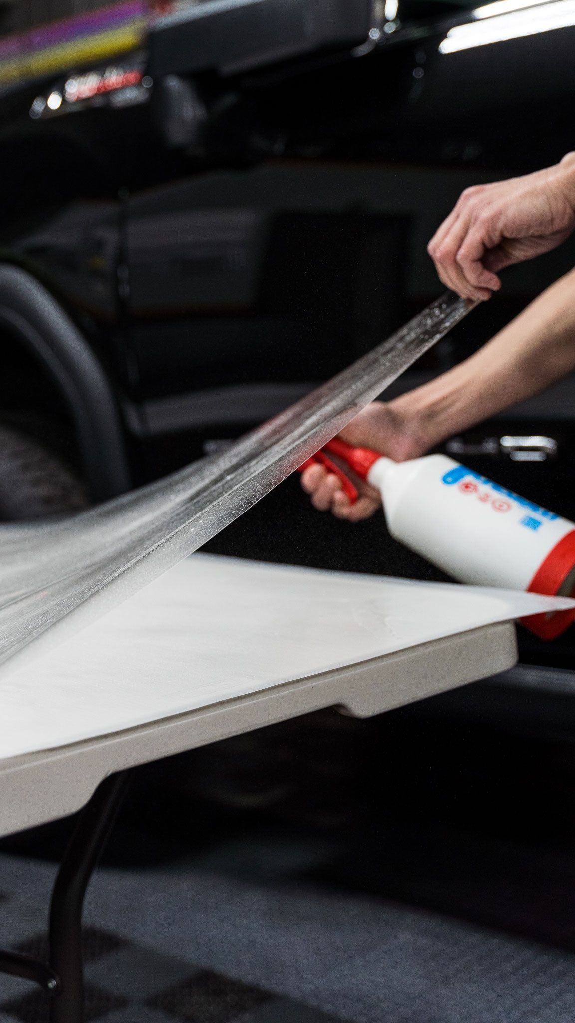 Person spraying a sheet of clear plastic on a white table next to a black vehicle.