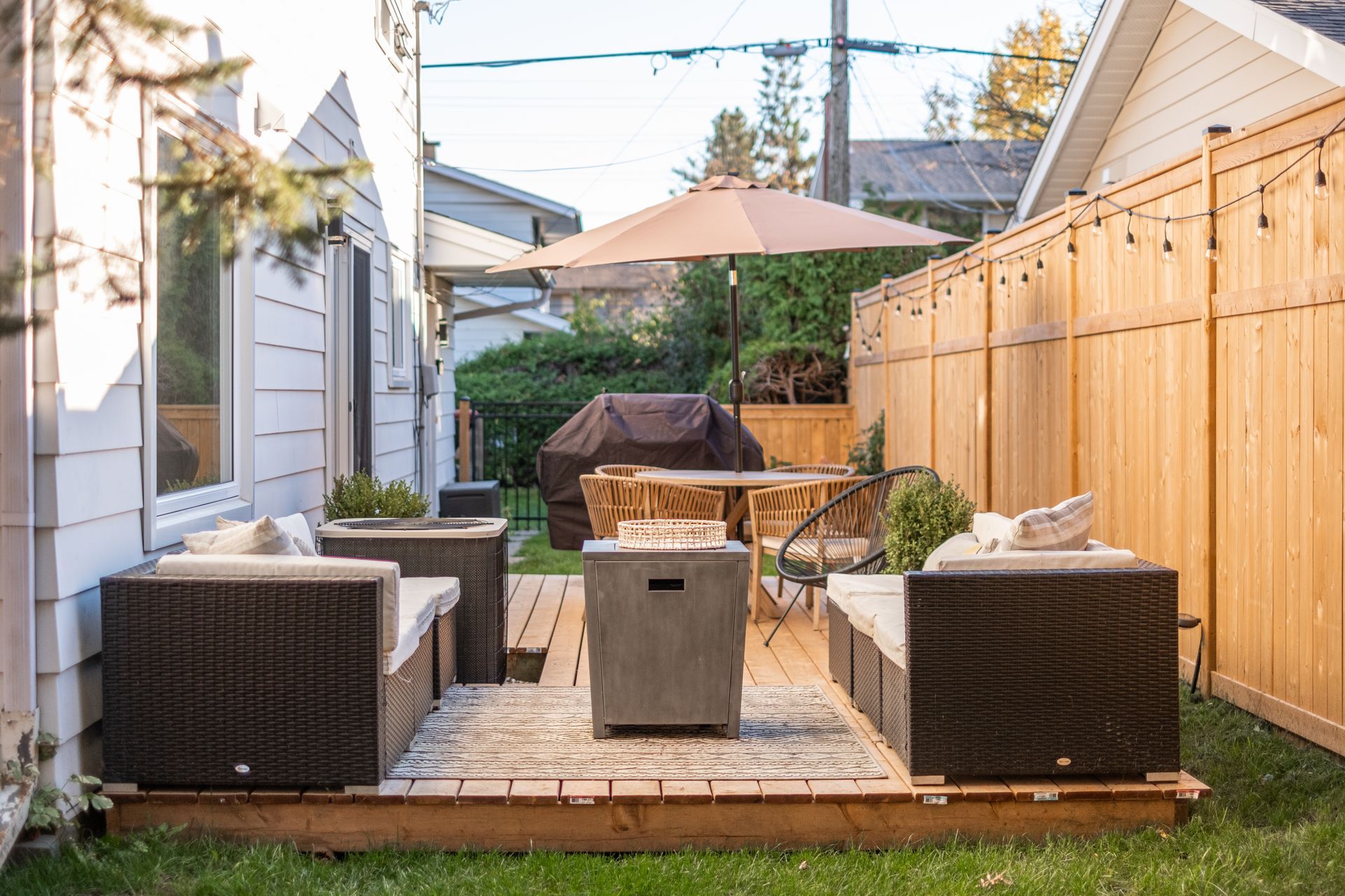 A backyard with a wooden deck , chairs , umbrella , table and grill.