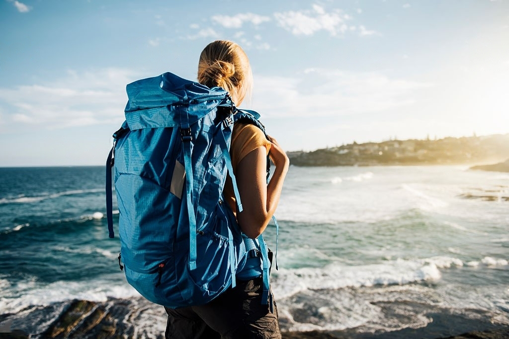 A woman with a blue backpack is standing on a rock near the ocean.