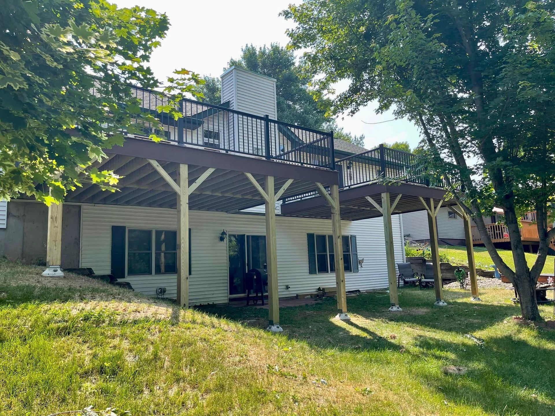 Two-story house with a raised deck and support posts, surrounded by trees and a grassy yard.