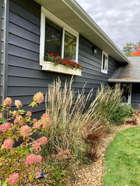 Gray house exterior with flower bed, pink blooms, and tall ornamental grass under cloudy sky