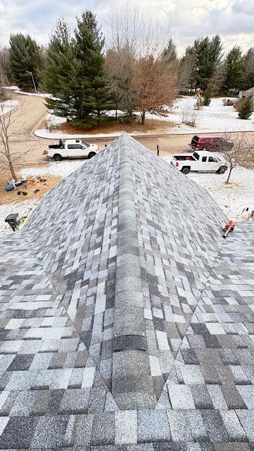 View from a gray shingled roof toward a snowy yard with evergreen trees and parked white trucks below