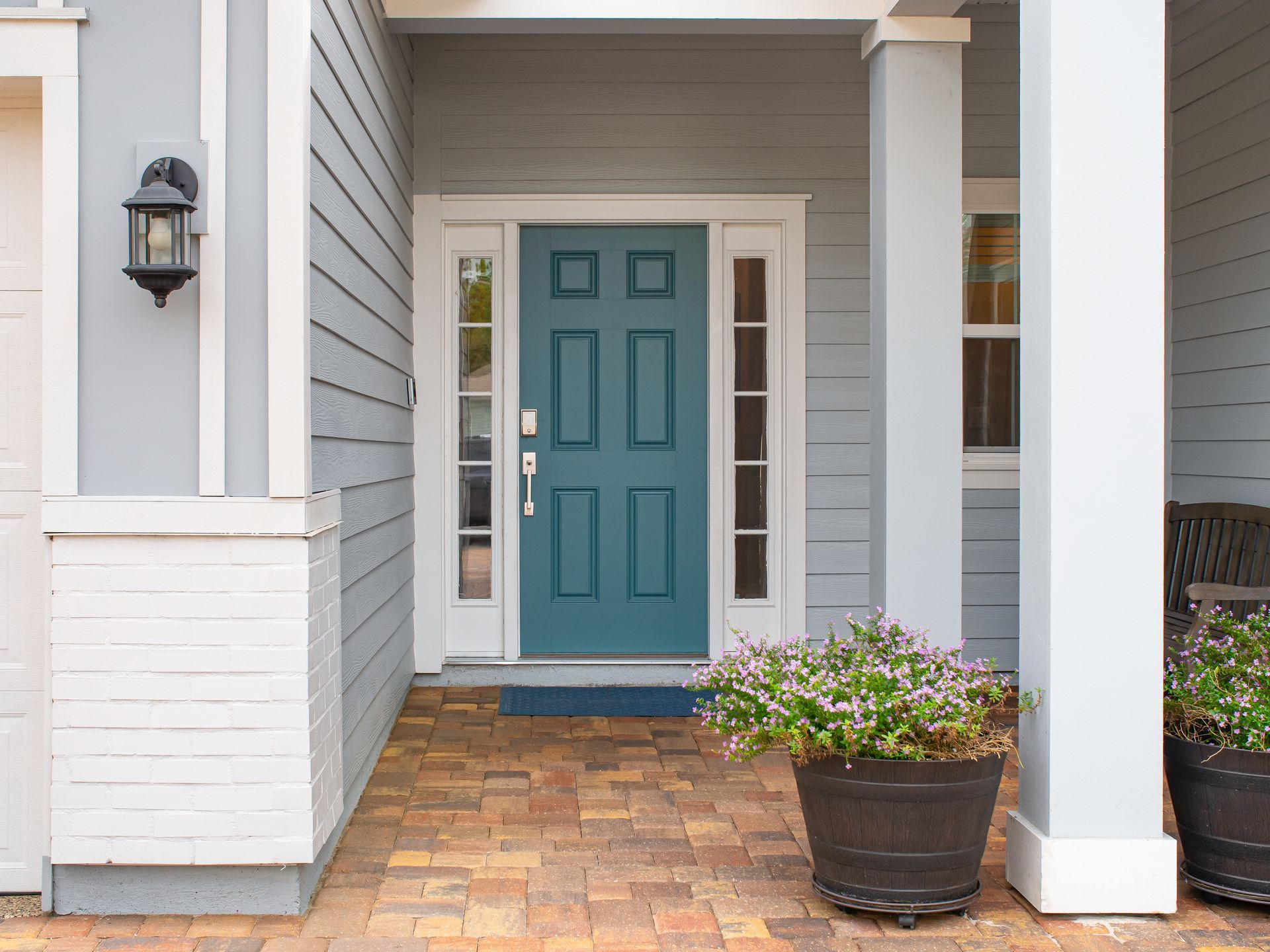 Front porch with a teal front door, white columns, gray siding, and a potted plant on brick pavers.