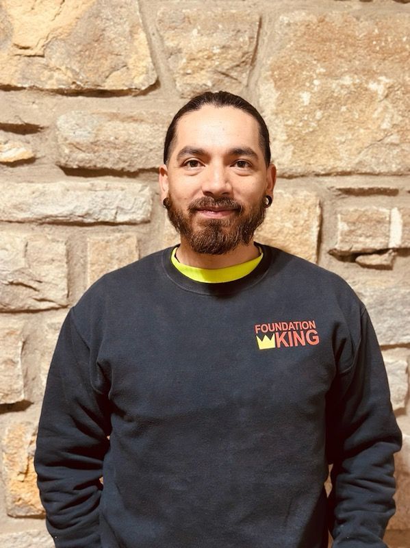 A man wearing a foundation king sweatshirt is standing in front of a stone wall.