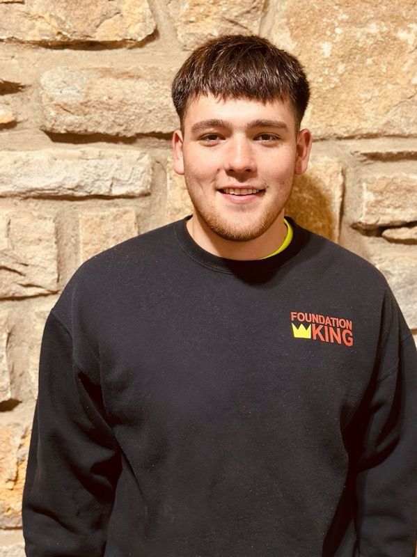 A young man wearing a black foundation king sweatshirt is standing in front of a stone wall.