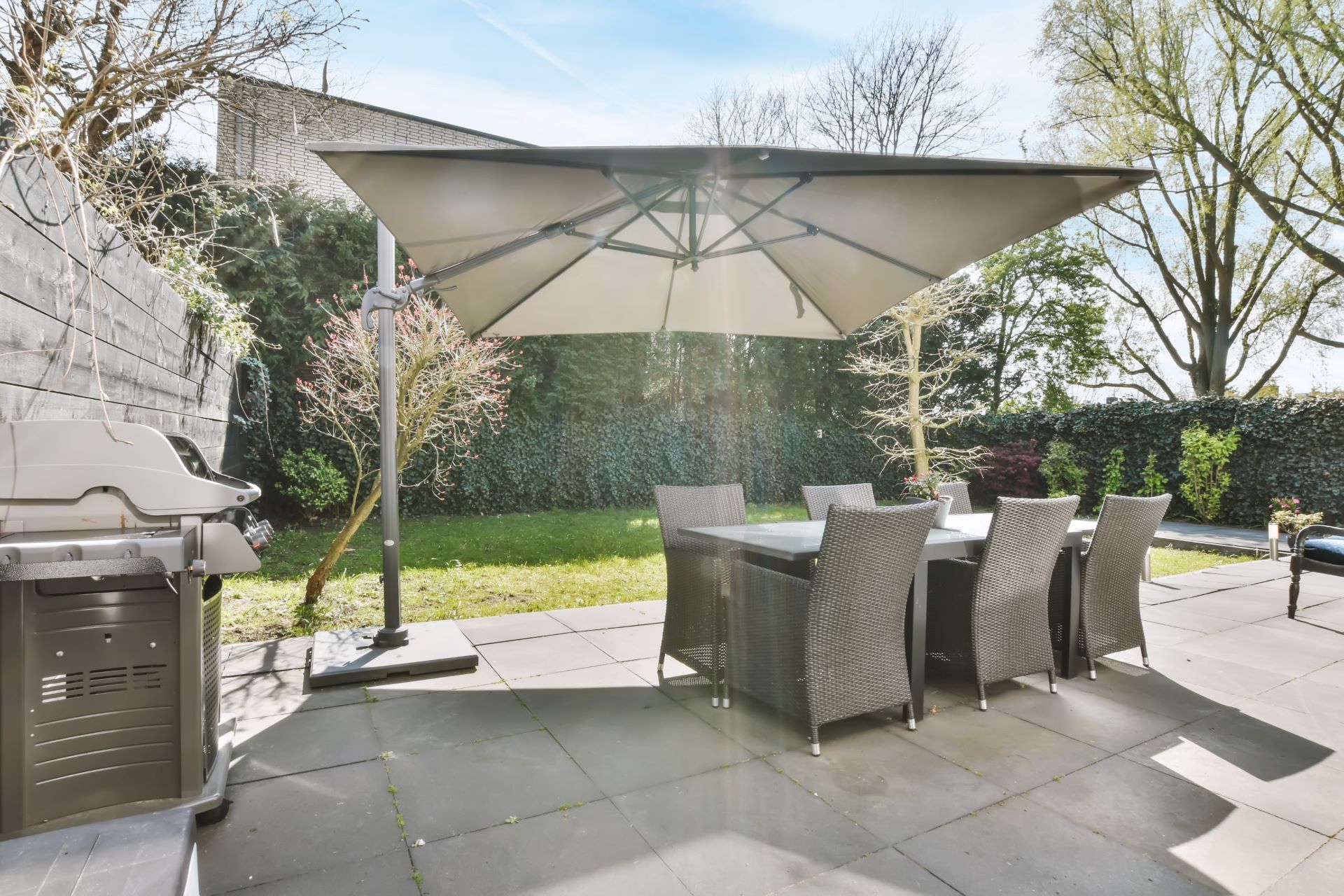 Patio with table and chairs under a large umbrella, grill on left, green lawn and trees in background.