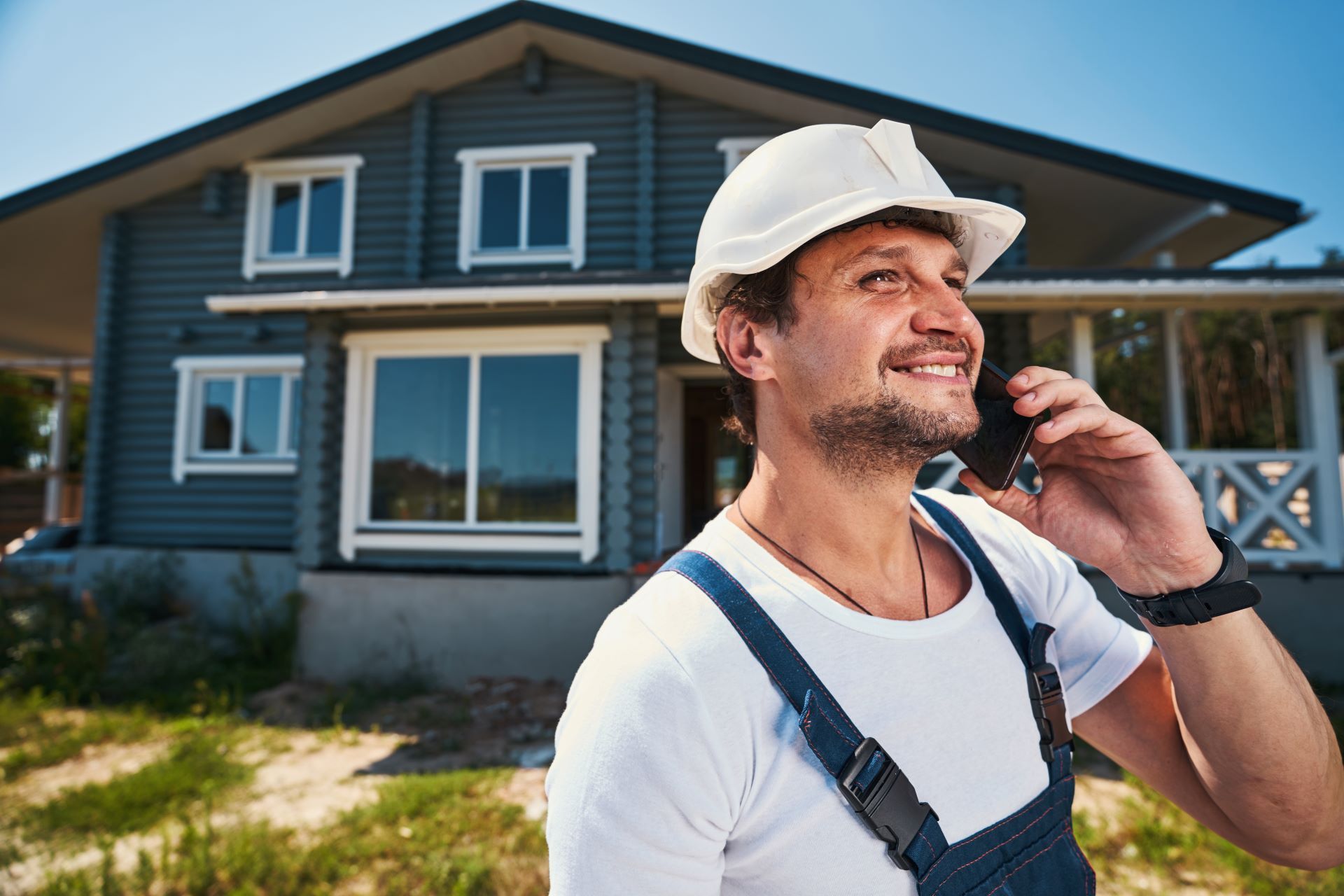 Construction worker in a white hard hat and blue overalls talking on a phone, in front of a blue house.