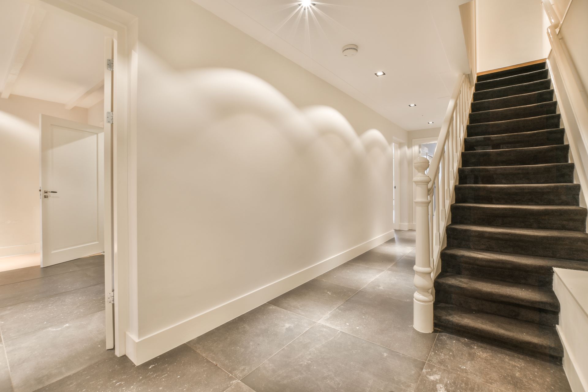 Hallway with stairs, a white wall, and dark gray tile floor. A door is visible on the left.