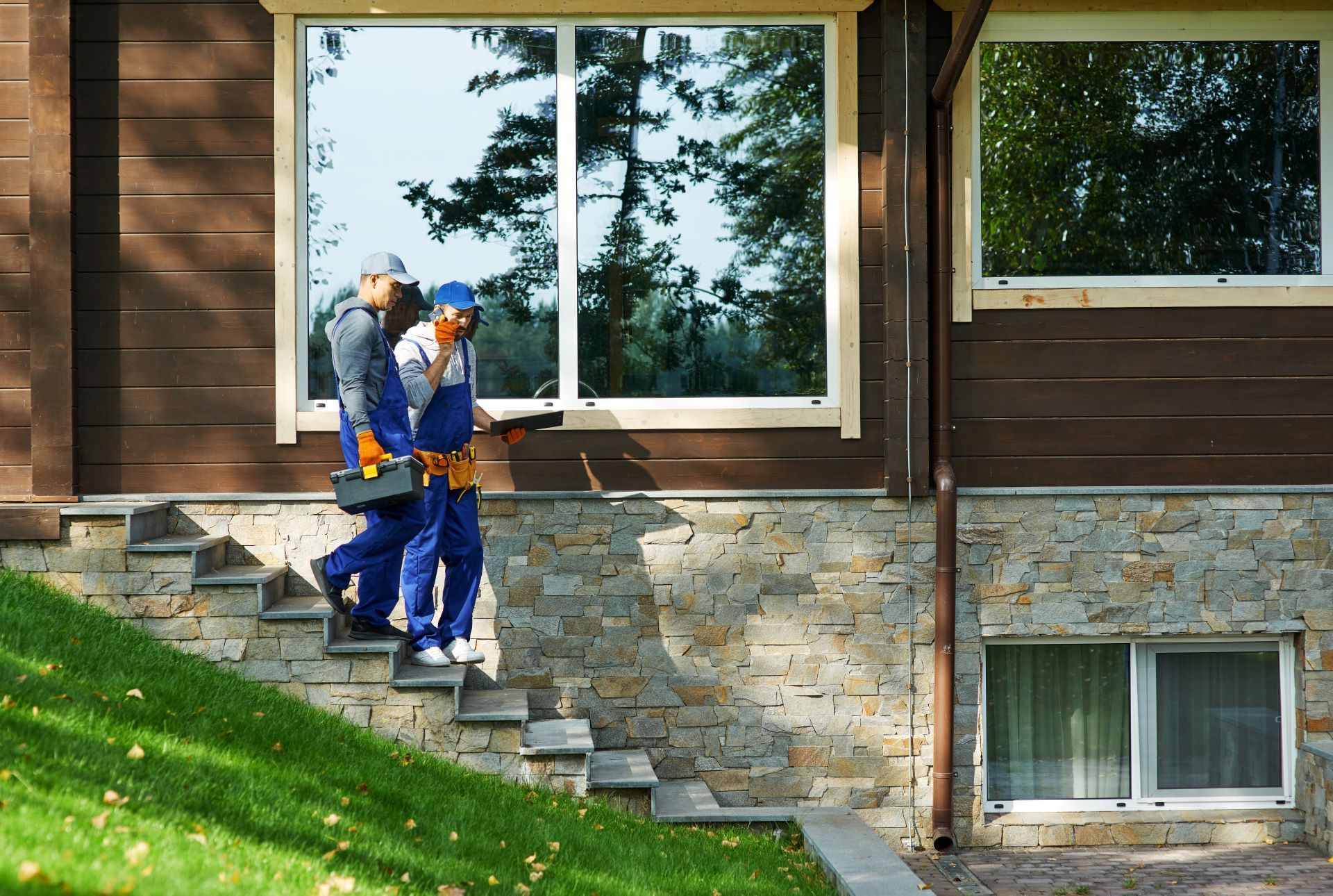 Two workers in blue overalls descend stone steps near a house with large windows.