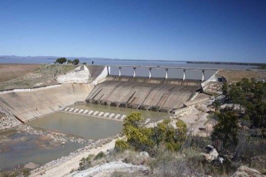 Dry dam spillway beside a reservoir, with concrete channels and sparse desert vegetation under a clear sky