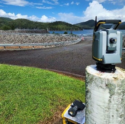 Surveying equipment on a concrete post beside a paved path, with ocean and green hills in the background