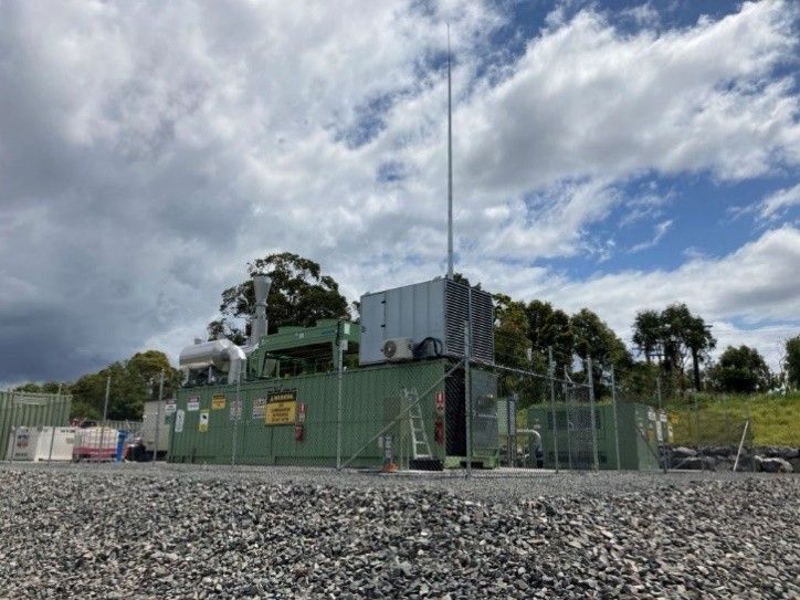 Green industrial building with tall antenna behind a chain-link fence under a cloudy sky
