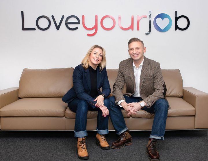 Julie Warner and Haydon Warner  sitting on a tan couch beneath a colorful “Loveyourjob” sign.