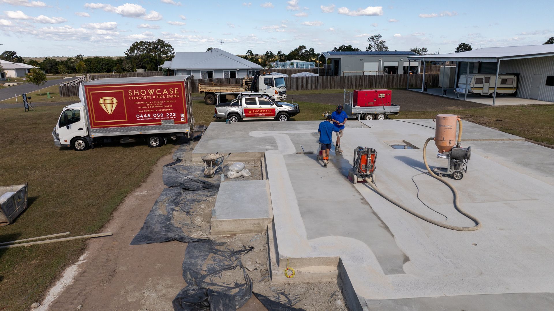 Construction workers using machinery on a concrete foundation in a residential area; red trucks and buildings visible. — Showcase Concrete & Polishing in Moore Park Beach, QLD