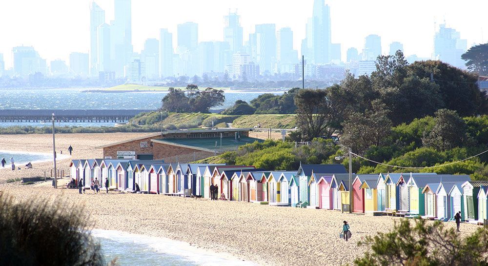 Colorful beach boxes line a sandy beach, with a city skyline in the background, near the water.