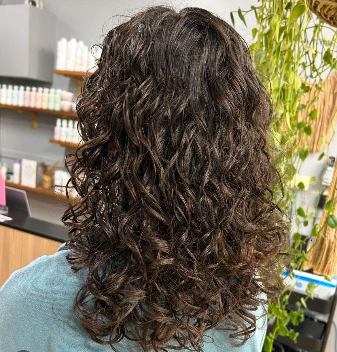 A Woman With Curly Hair Is Standing In Front Of A Shelf — M & Co Hair Salon In Atherton, QLD