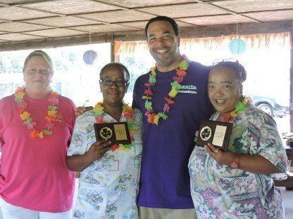 Four people wearing leis hold small awards indoors under a thatched roof.