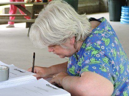 Person in a blue floral shirt writing on paper at a table outdoors.