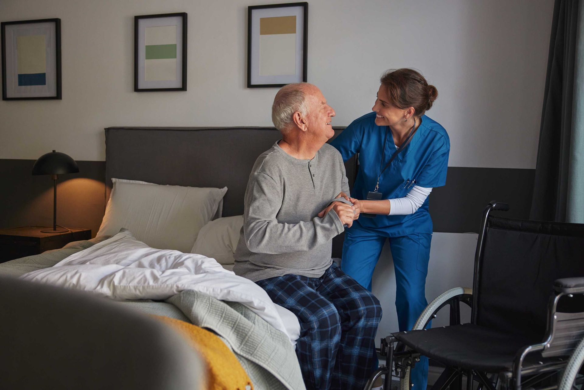 Caregiver in blue helping an elderly person sit on a bed in a bedroom