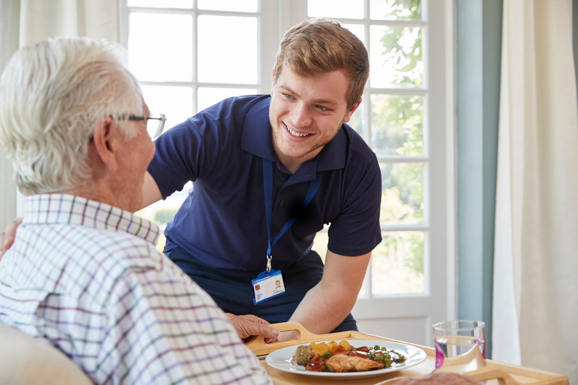 Caregiver smiling at an older man seated by a meal in a bright dining room