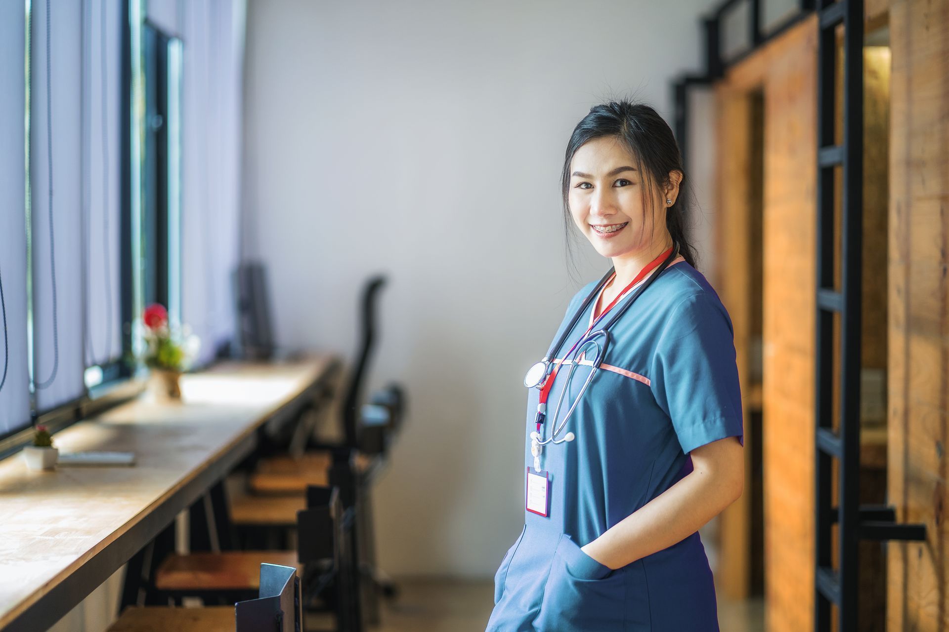 Nurse in blue scrubs standing in a bright hospital hallway, smiling beside a window and chairs