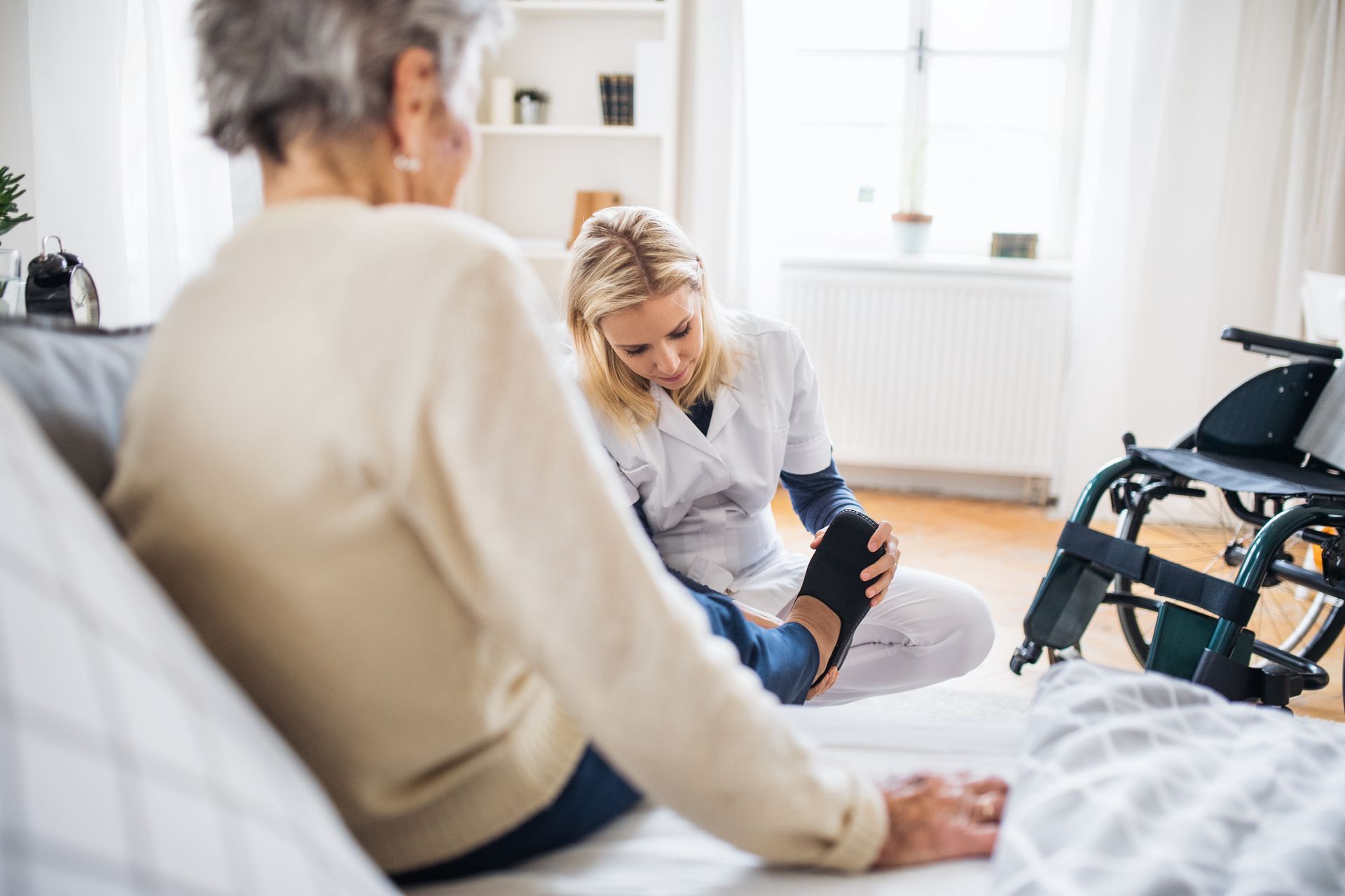Caregiver helping a patient stretch in a bright home room, with a wheelchair nearby.