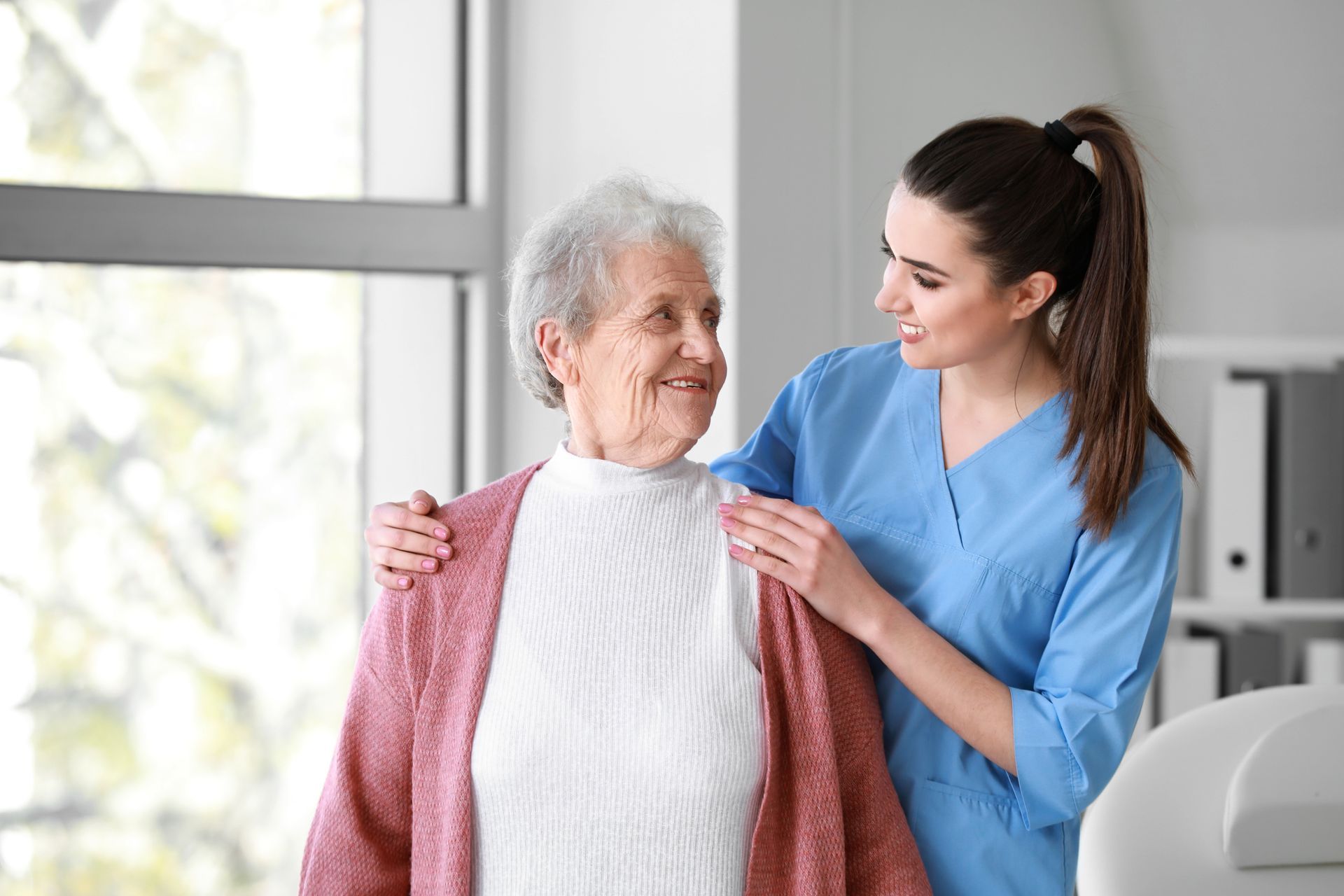 Caregiver in blue scrubs helps an elderly woman wear a pink cardigan in a bright room.
