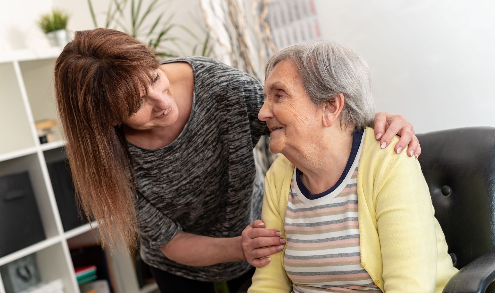 Woman comforting an elderly seated person indoors, smiling and leaning in beside a couch