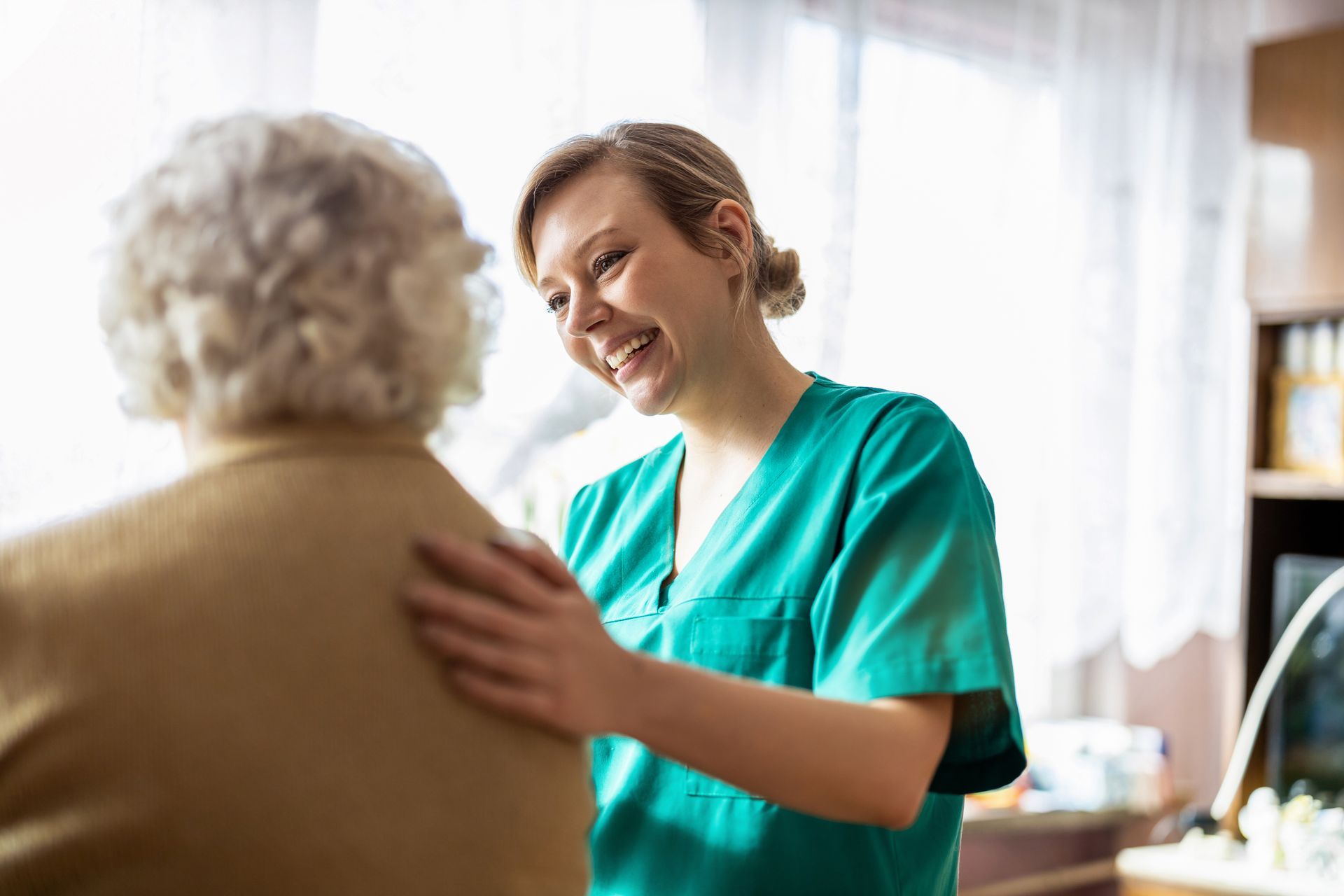 Nurse in teal scrubs gently comforts an elderly patient in a bright clinic room