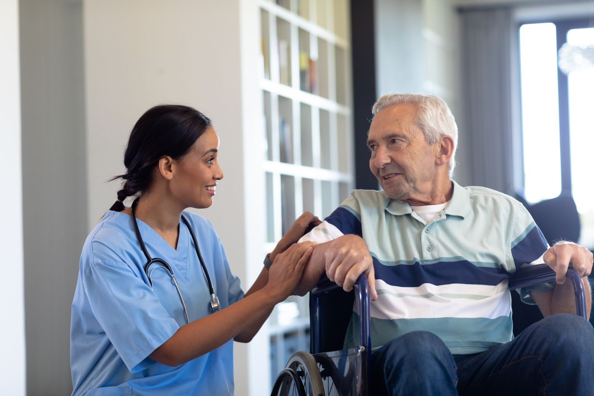 Nurse in blue scrubs gently talks with a seated patient in a bright clinic hallway.