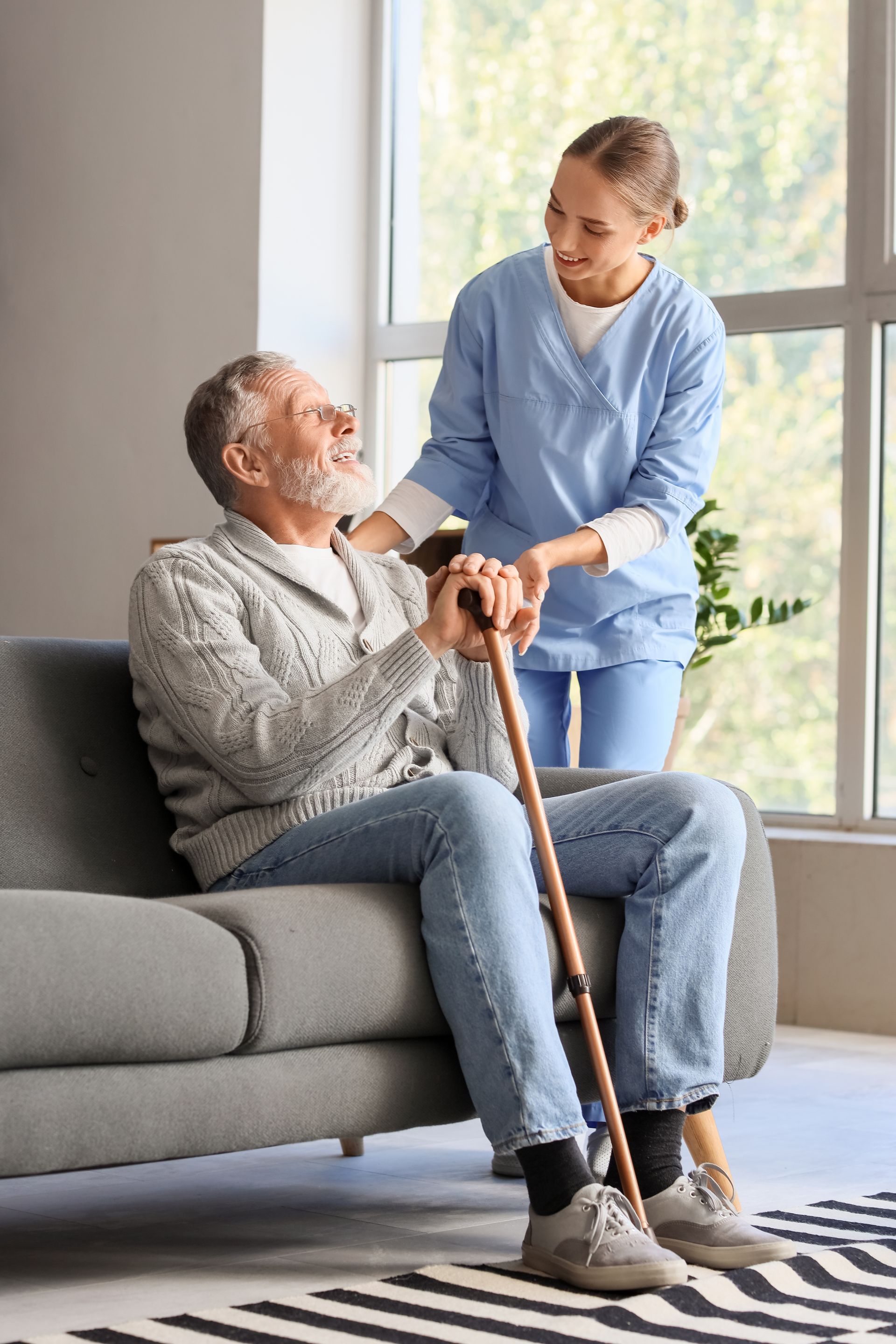Caregiver helping a seated man with a cane on a sofa in a bright living room