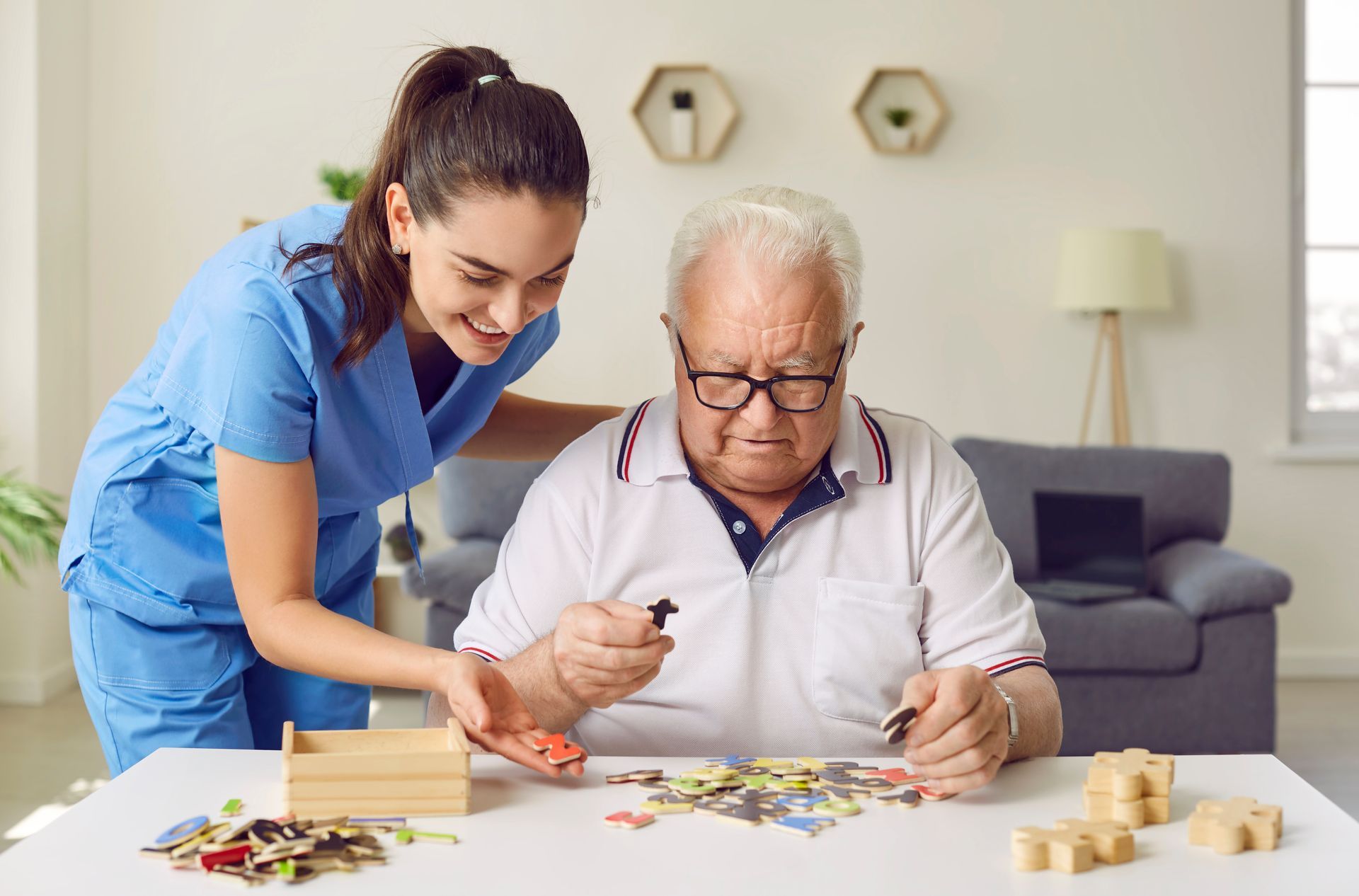 Caregiver assisting an older man with a tabletop puzzle in a bright living room