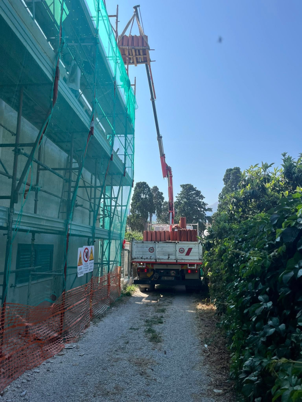 Un'autogru solleva una struttura in legno accanto a un edificio coperto da impalcature e reti, lungo una stretta strada sterrata.