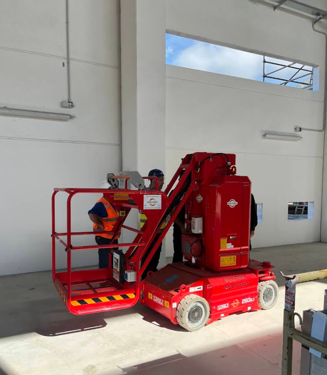 Piattaforma aerea rossa con operai all'interno di un magazzino. Un operaio indossa un giubbotto di sicurezza.