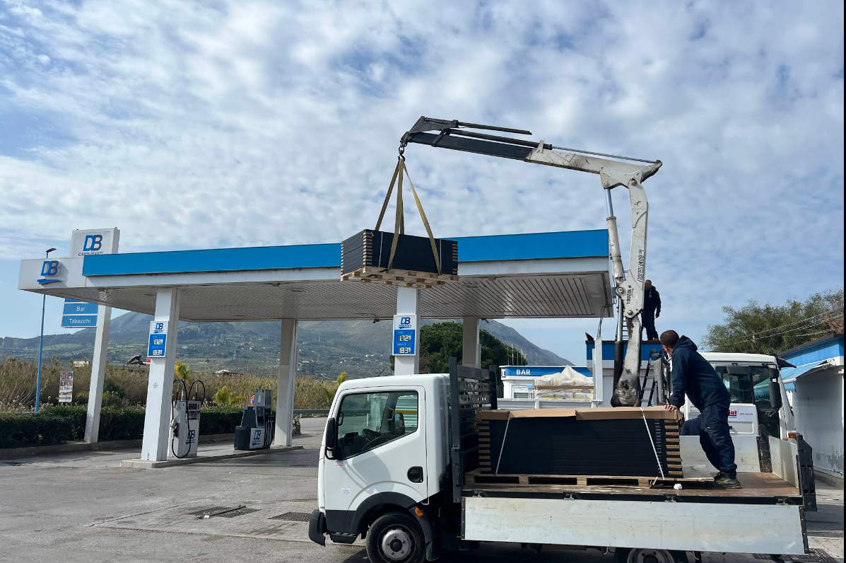 Una gru montata su un camion solleva un oggetto rettangolare scuro in una stazione di servizio. Una persona sul camion. Cielo visibile.