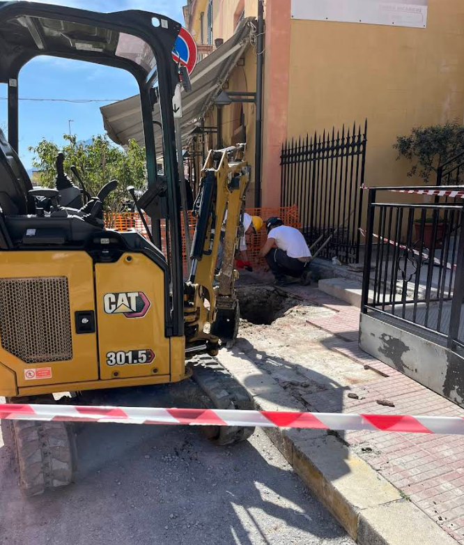 Escavatore giallo CAT 301.5 e operaio che scavano vicino a un edificio con nastro di avvertenza rosso/bianco.