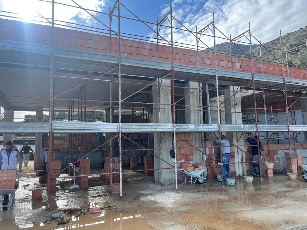 Interno di un cantiere; colonne di cemento sostengono un pavimento, muri di mattoni in costruzione, due operai.