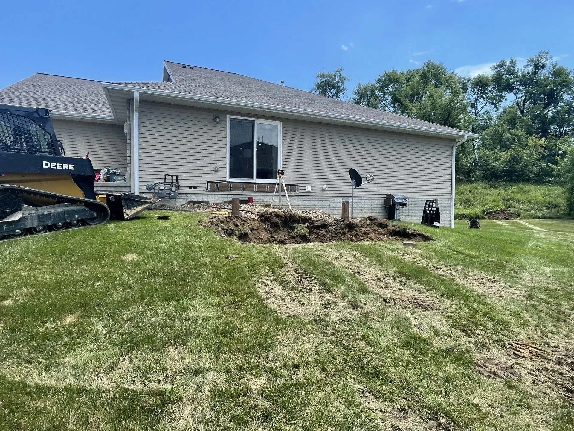 A bulldozer digs dirt near a house with a sliding glass door; grass and trees in the background.