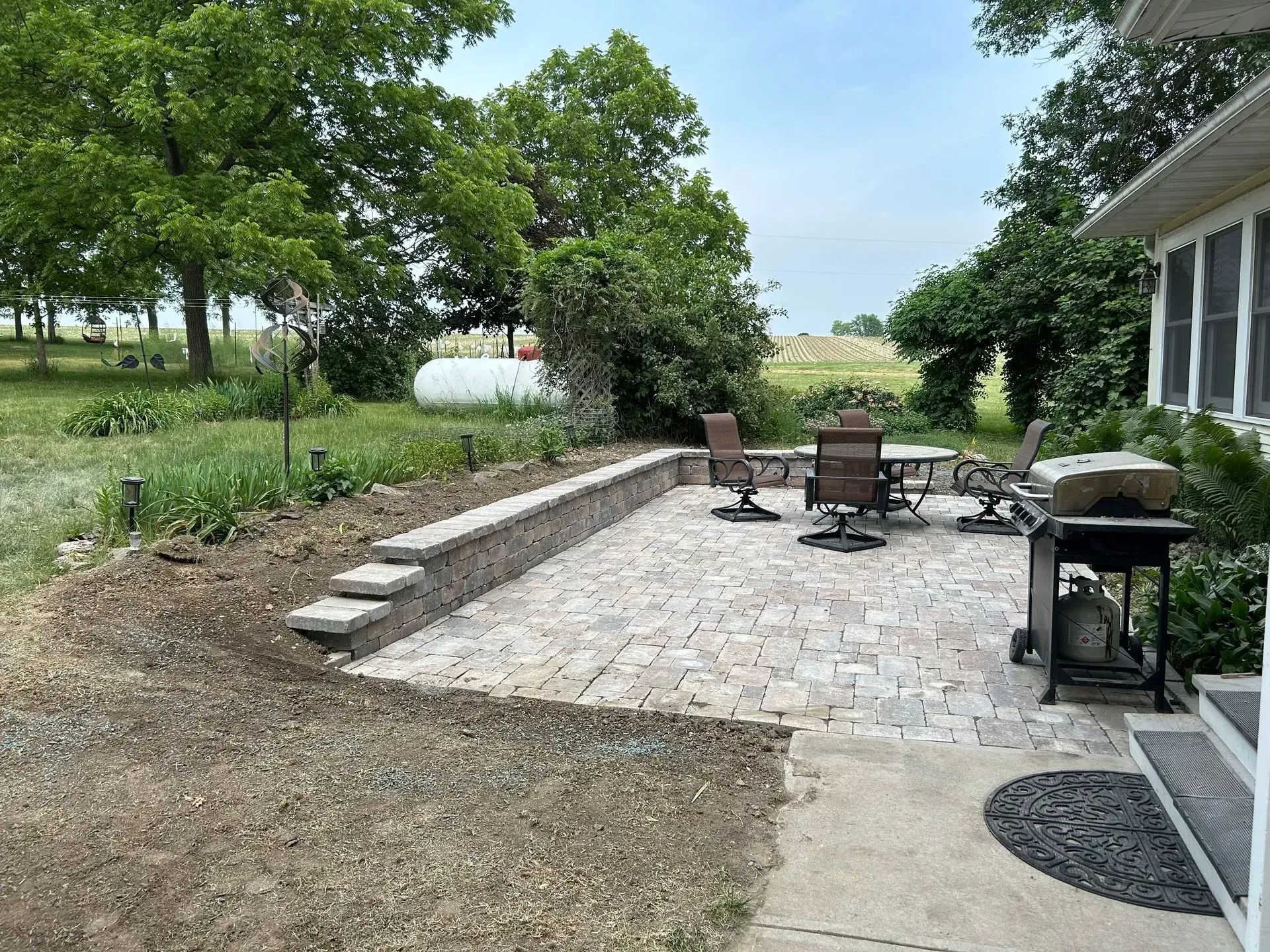 Paver patio with retaining wall and steps, featuring outdoor furniture and a grill, in a backyard setting.