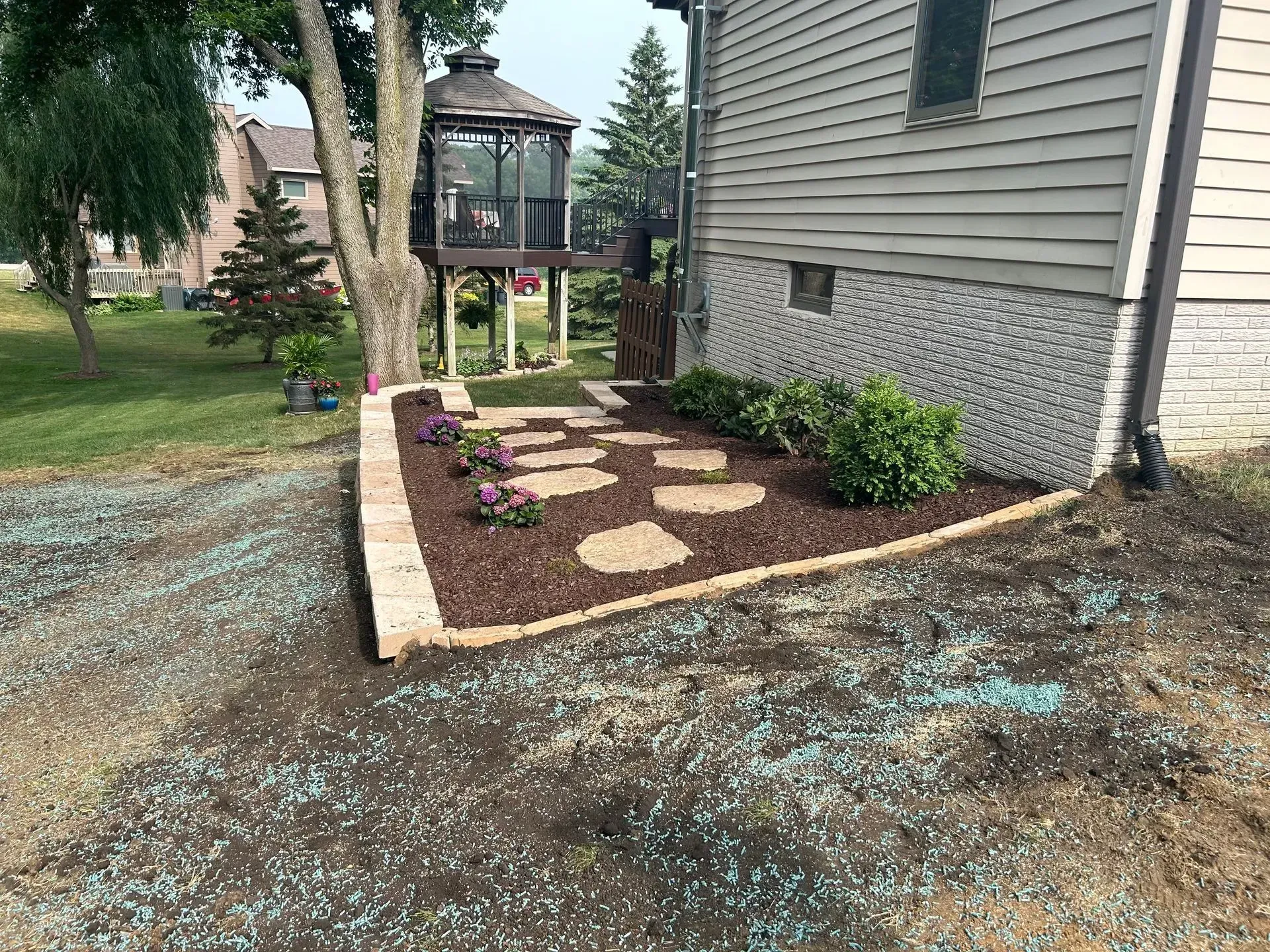Landscaped garden bed with stone path next to a house, featuring flowers and mulch, surrounded by grass.