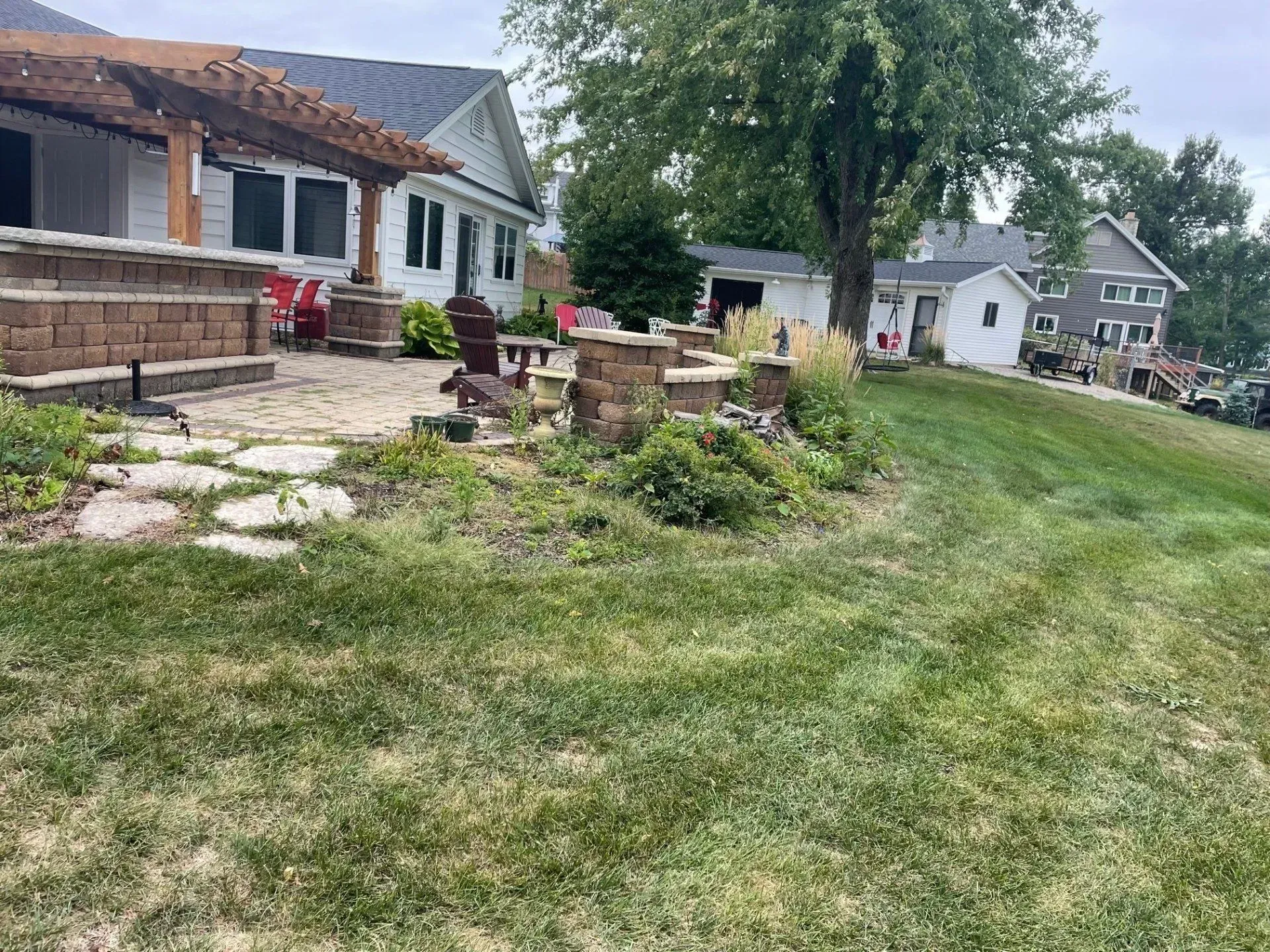 Lakeside backyard with patio, stone walkway, and lawn. Houses in the background under a cloudy sky.